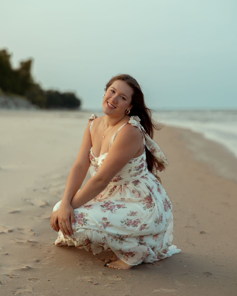 A person in a floral dress sits playfully on a sandy beach at sunset with waves in the background.