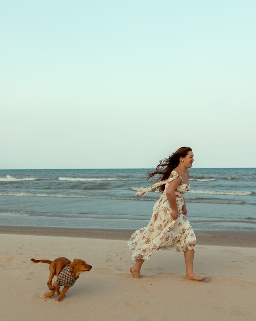 A person and a dog run playfully along a serene beach at sunset, their silhouettes casting long shadows on the sand.
