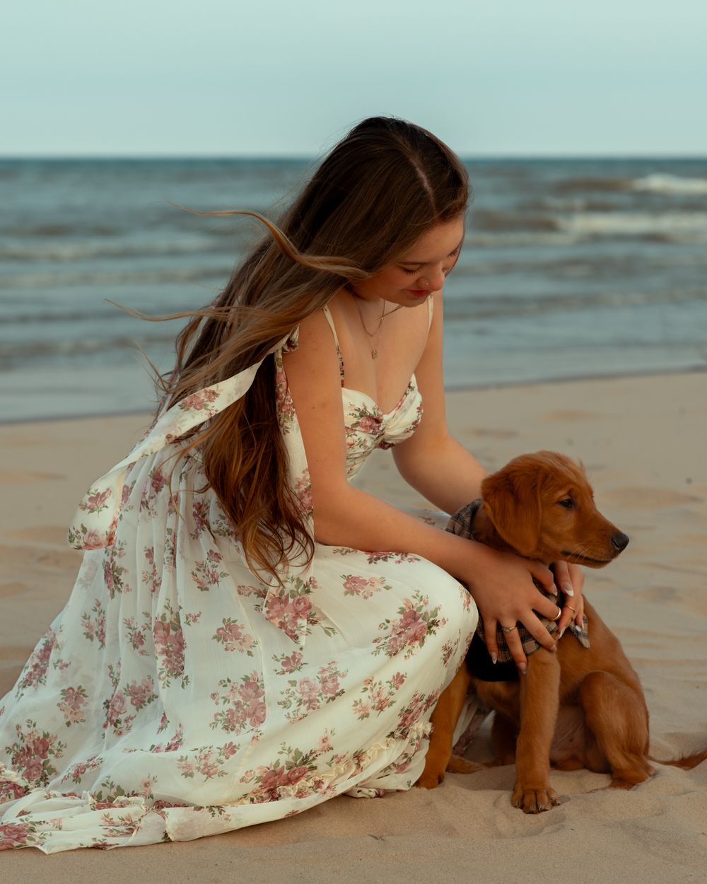 A person in a floral dress sits on the beach with a brown puppy as waves crash in the background.