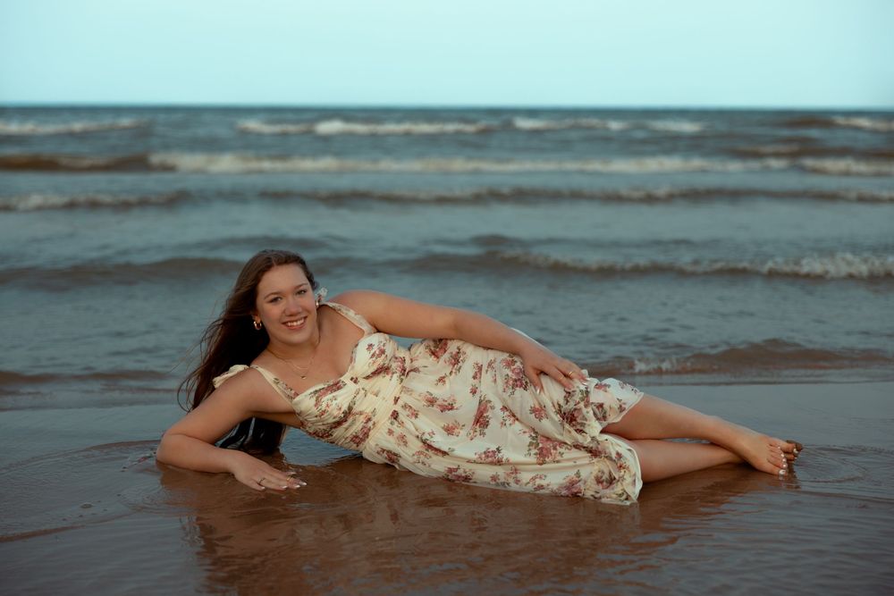 A person in a floral dress poses on the beach at sunset with gentle waves in the background.