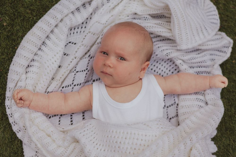 a baby frowns, laying on the grass in a white vest, wrapped in a white blanket