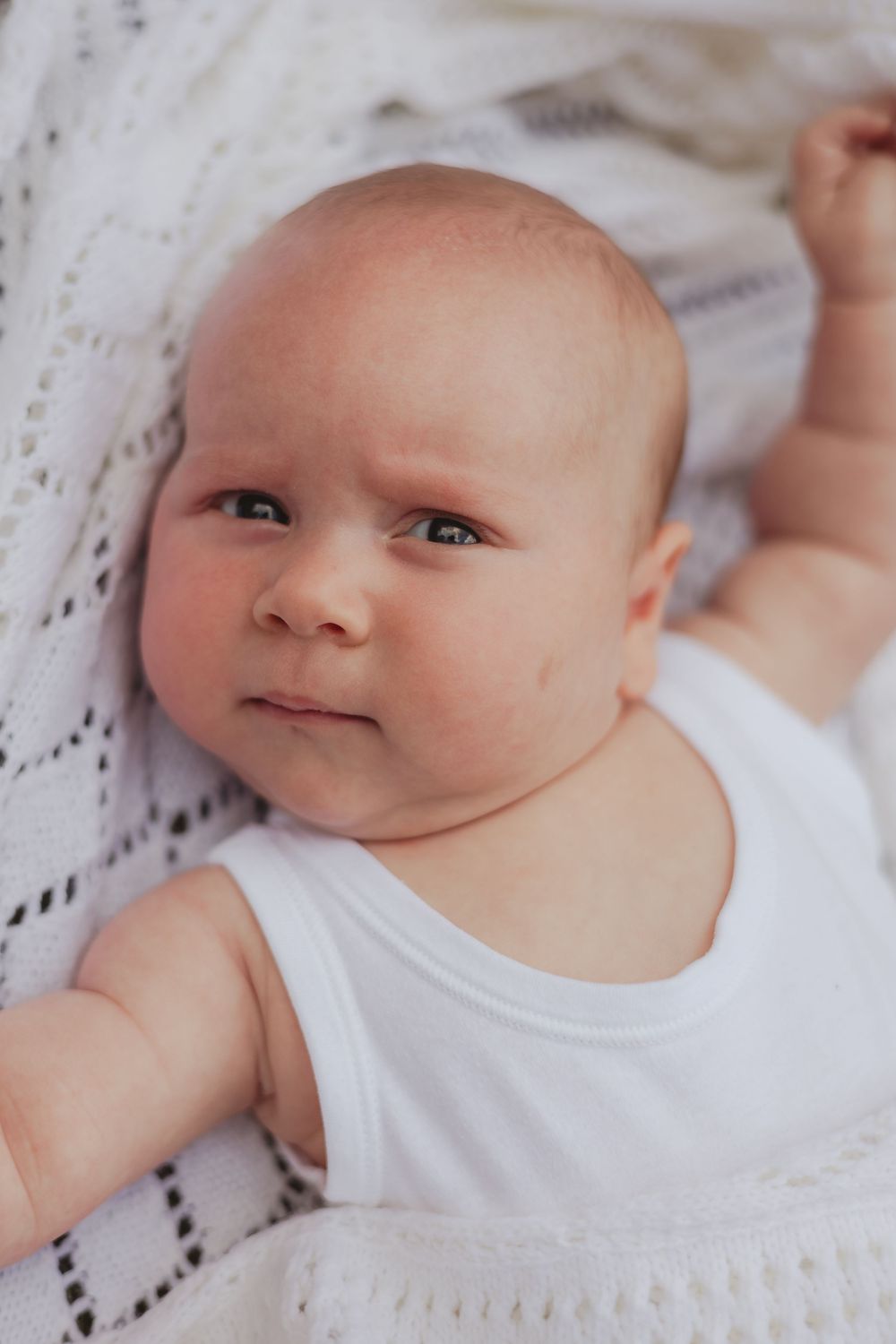 a baby frowns, laying on the grass in a white vest, wrapped in a white blanket