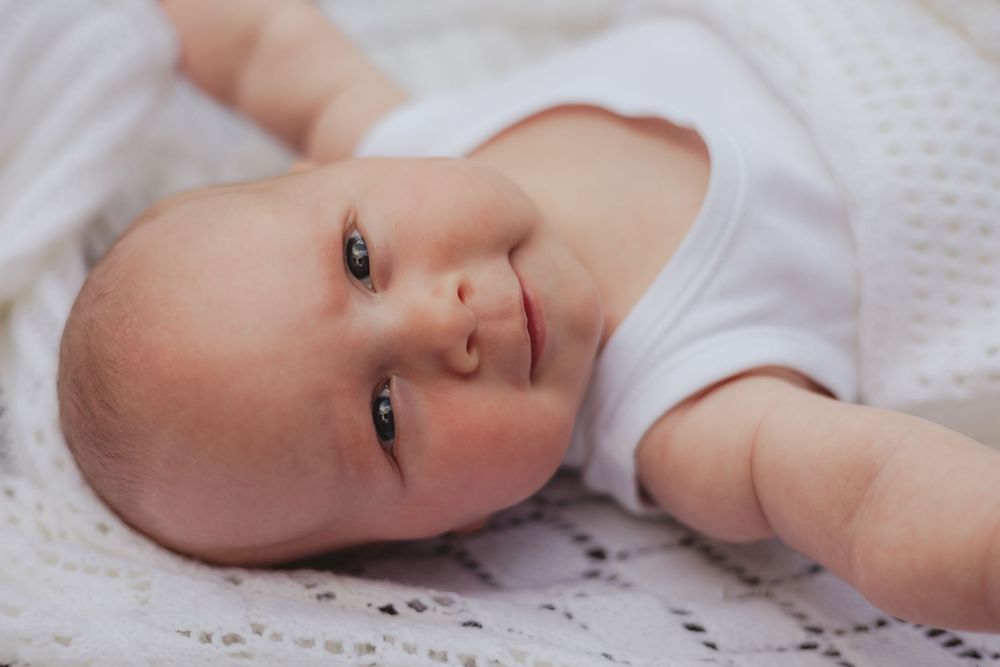 a baby smirks, laying on the grass in a white vest and blanket