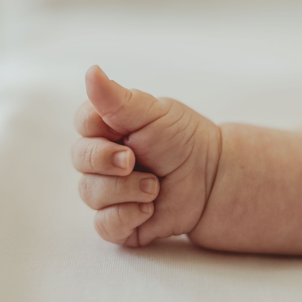 A delicate image of a small baby's hand, showing tiny details of fingernails and creases. The hand is clasped in a thumbs up pose.