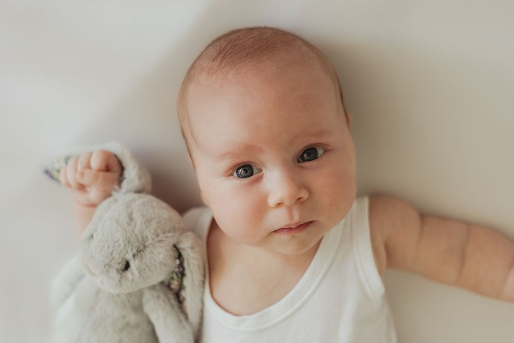 A baby in a white vest, clutching a grey bunny teddy, looks at the camera with big blue eyes