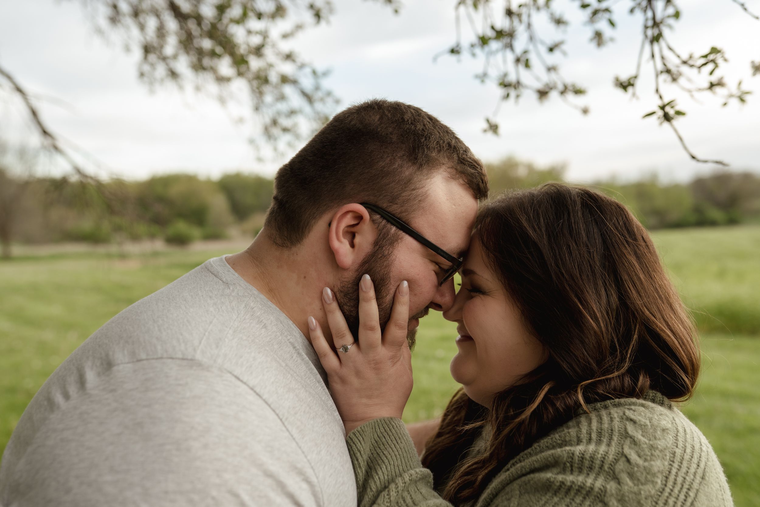 A couple embraces tenderly against a blurred outdoor background with trees and grass.