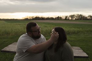 Two people share a tender moment on a wooden platform in a pastoral landscape at dusk.