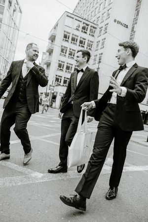 Three groomsmen in formal suits share a lighthearted moment on a city street corner.
