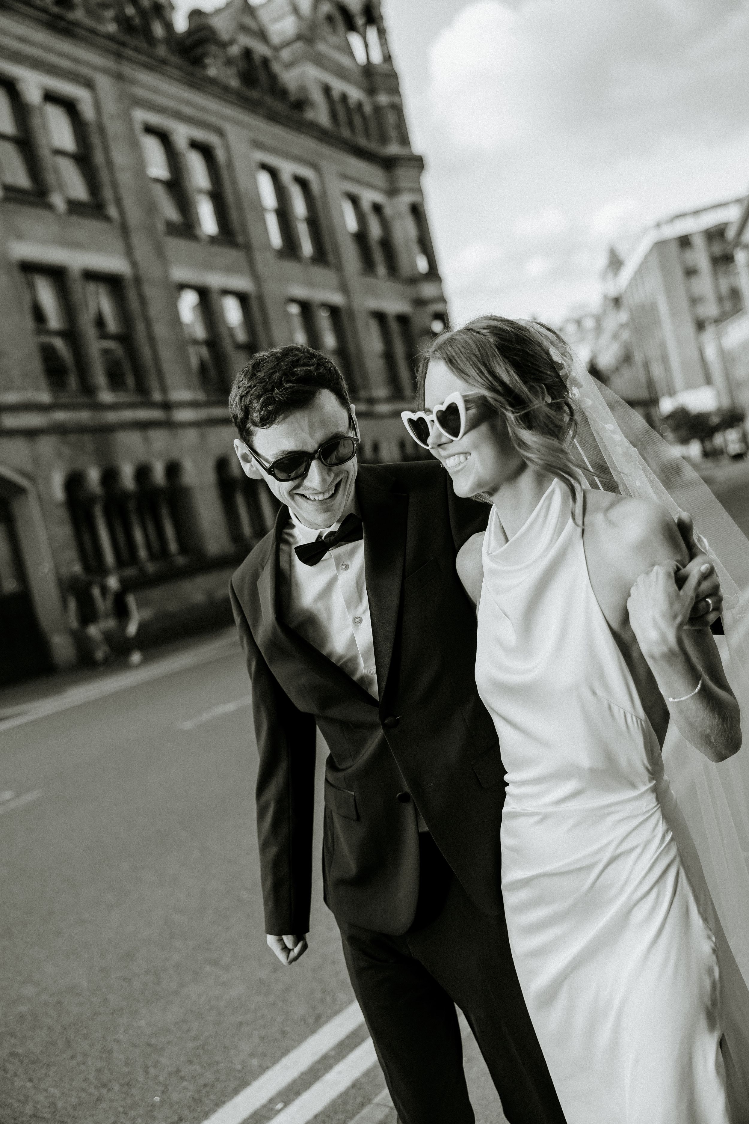 Black and white wedding photo of couple walking playfully on city street with historic buildings behind them.