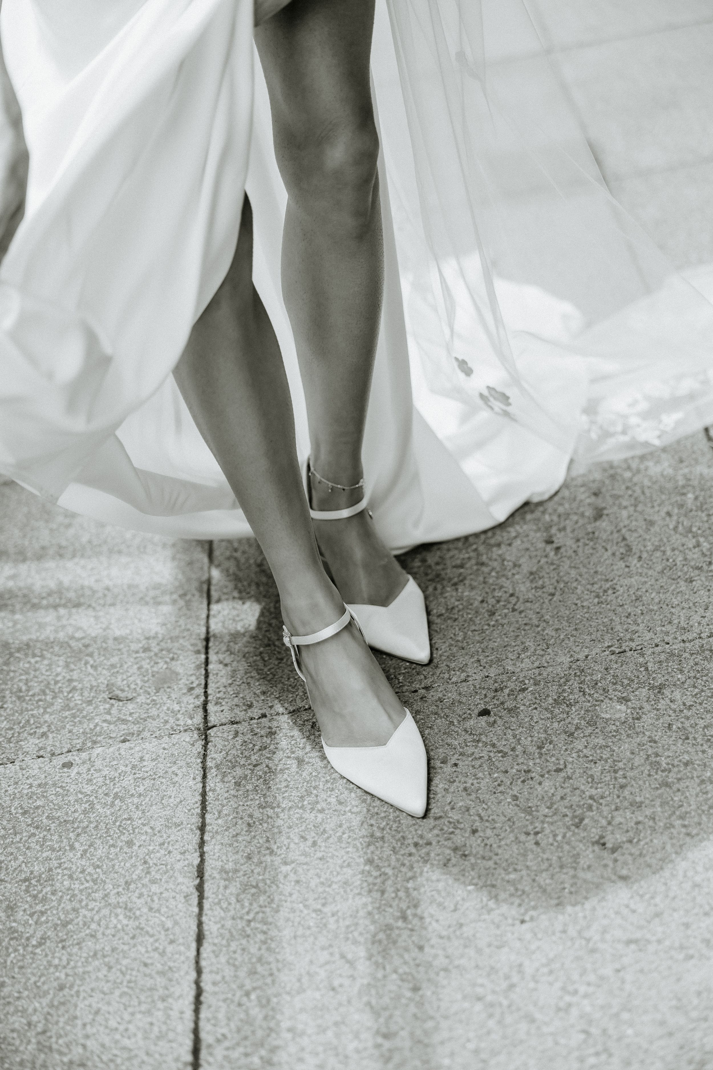 Black and white photograph showing elegant bridal shoes and flowing white dress hem on concrete ground.