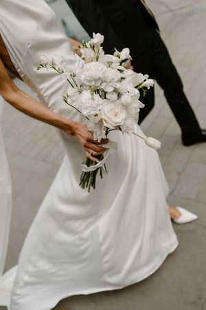 A bride in a flowing white dress holds an elegant bouquet of white roses and delicate florals against a gray floor surface.