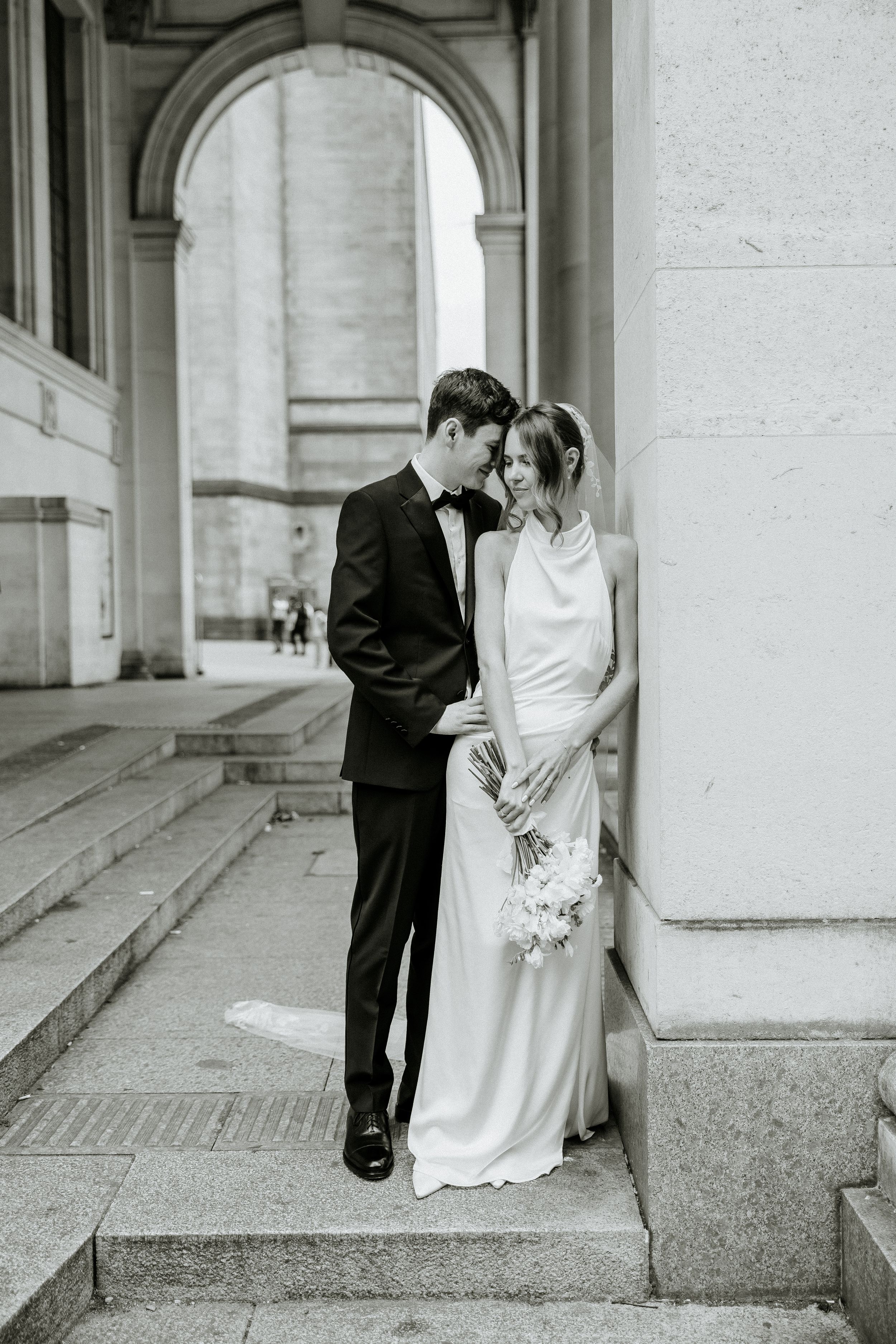Black and white wedding photo of a couple in elegant attire sharing an intimate moment under a classical archway.