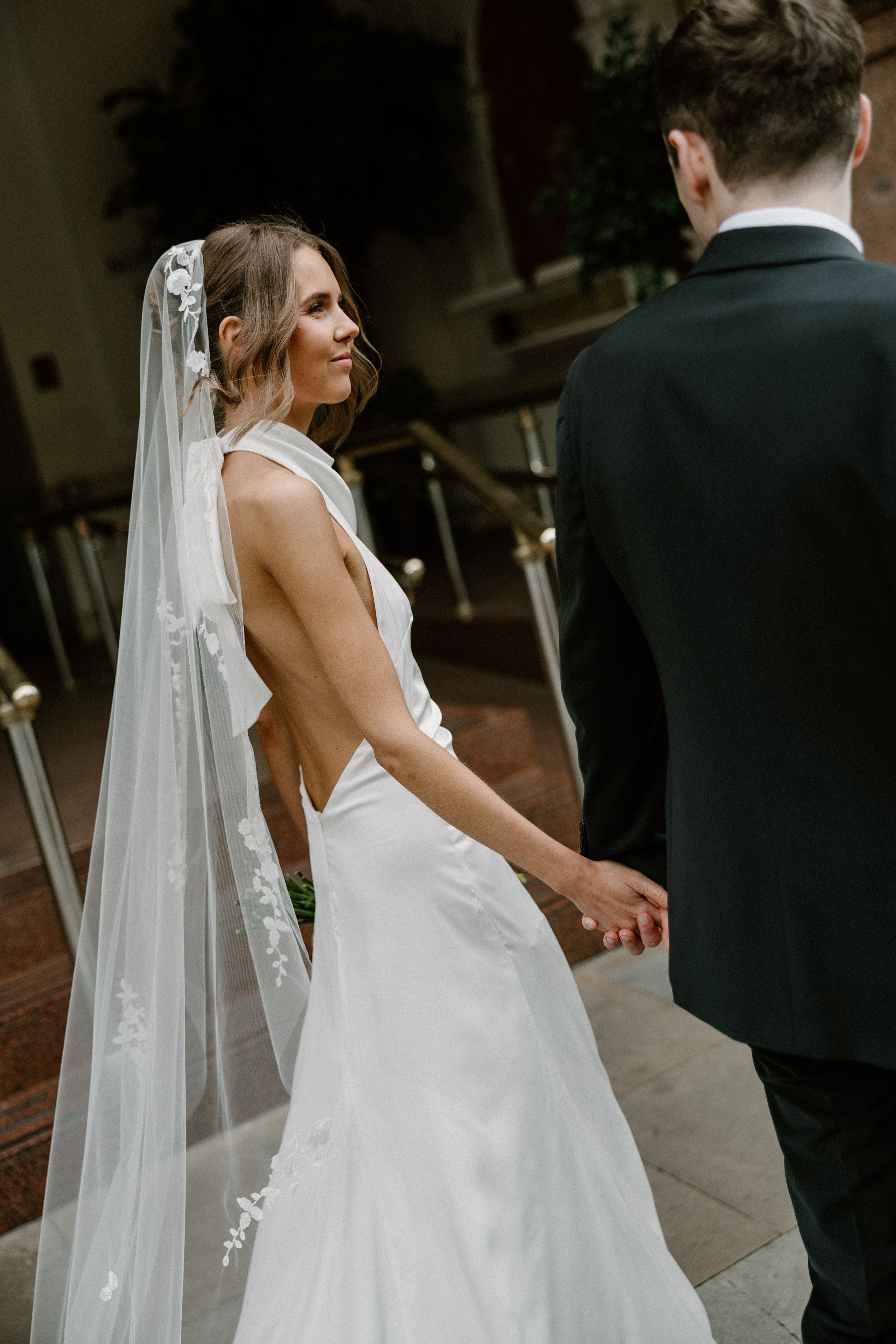 A bride in an elegant backless white wedding gown and long veil glances over her shoulder.