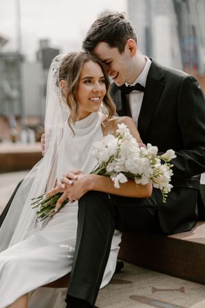 Elegant bride in white with flowing veil and groom in black share tender moments against urban backdrop.
