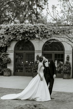 Black and white photo of a couple in wedding attire walking through a garden with arched windows.