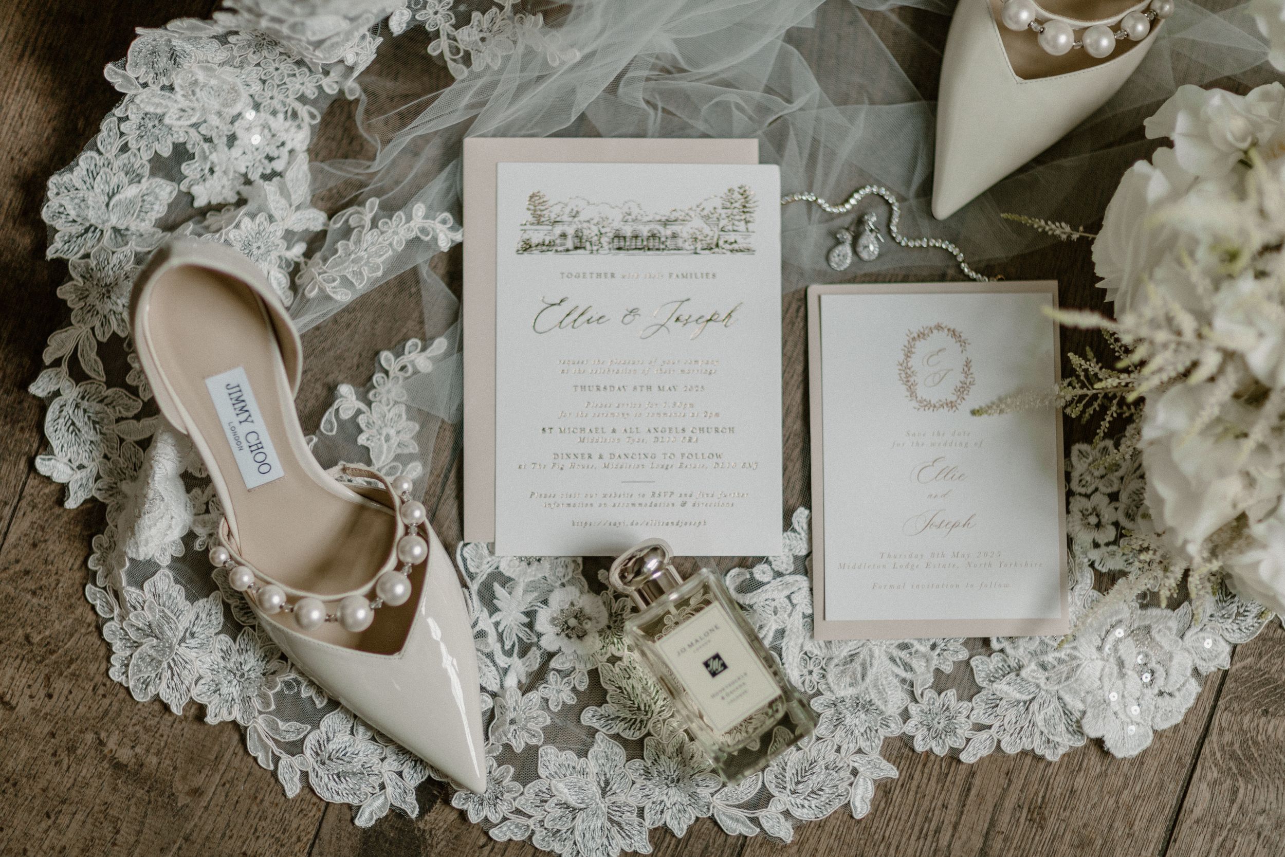 Wedding details flatlay with delicate white lace, pearls, invitations and floral elements on a neutral background.