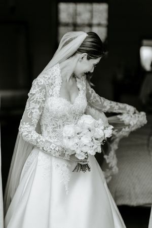 A bride in a long-sleeved lace wedding gown smiles while holding a bouquet of white roses.
