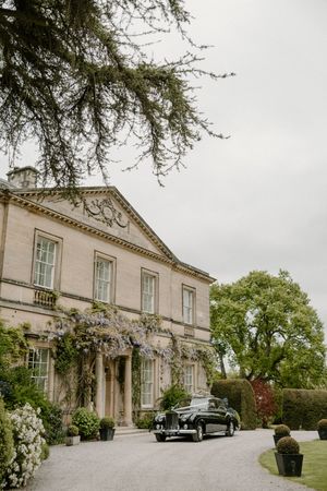 Classic vintage car parked in front of an elegant Georgian mansion with climbing wisteria and manicured gardens.