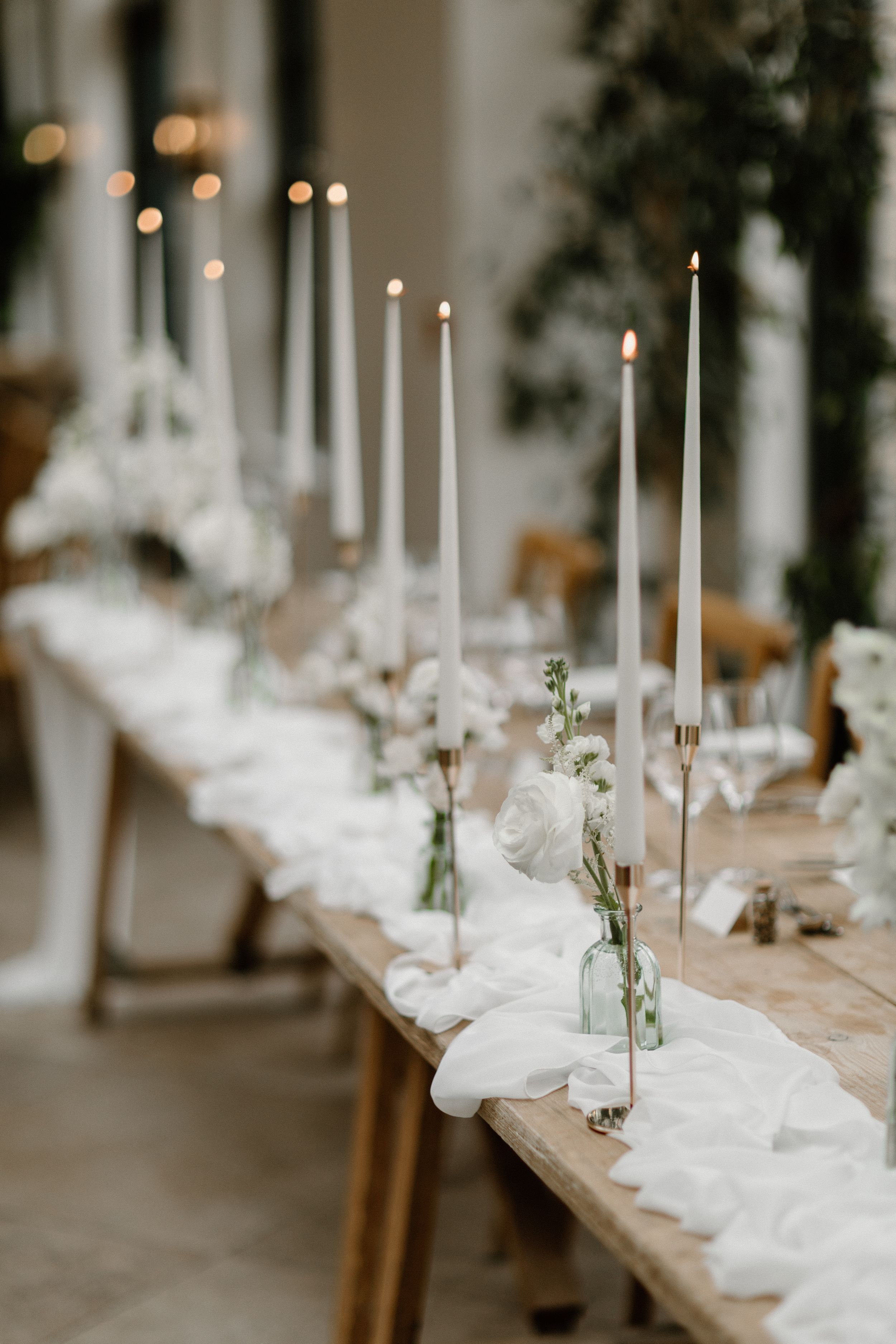 Long table setting with white linens, tall white candles, and small floral arrangements creating an elegant wedding reception ambiance.
