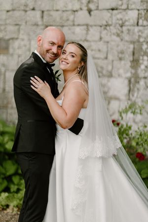 A couple embraces against a stone wall backdrop on their wedding day, with the bride wearing a flowing veil.
