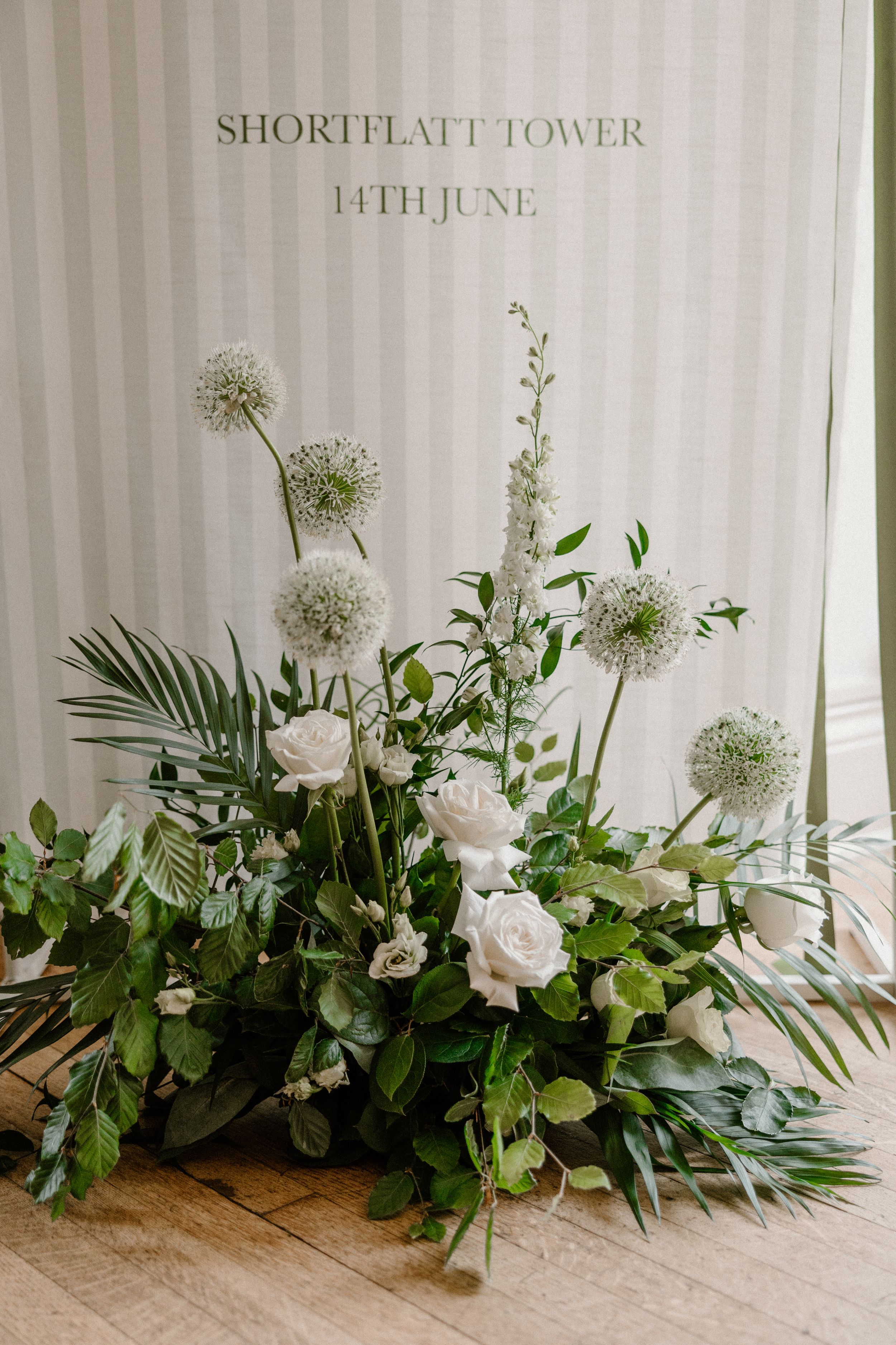 Elegant white floral arrangement with roses, allium flowers and palm leaves on a wooden table against a striped wall.
