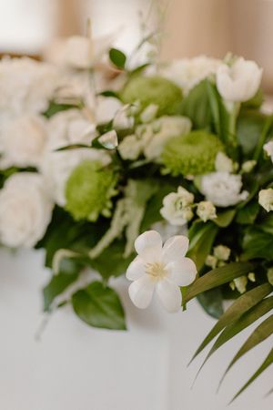 Close-up of elegant white wedding floral arrangement with green accents and foliage against a soft beige background.