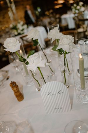 Elegant white roses in glass vases decorate wedding reception tables with delicate place cards and candlelight.