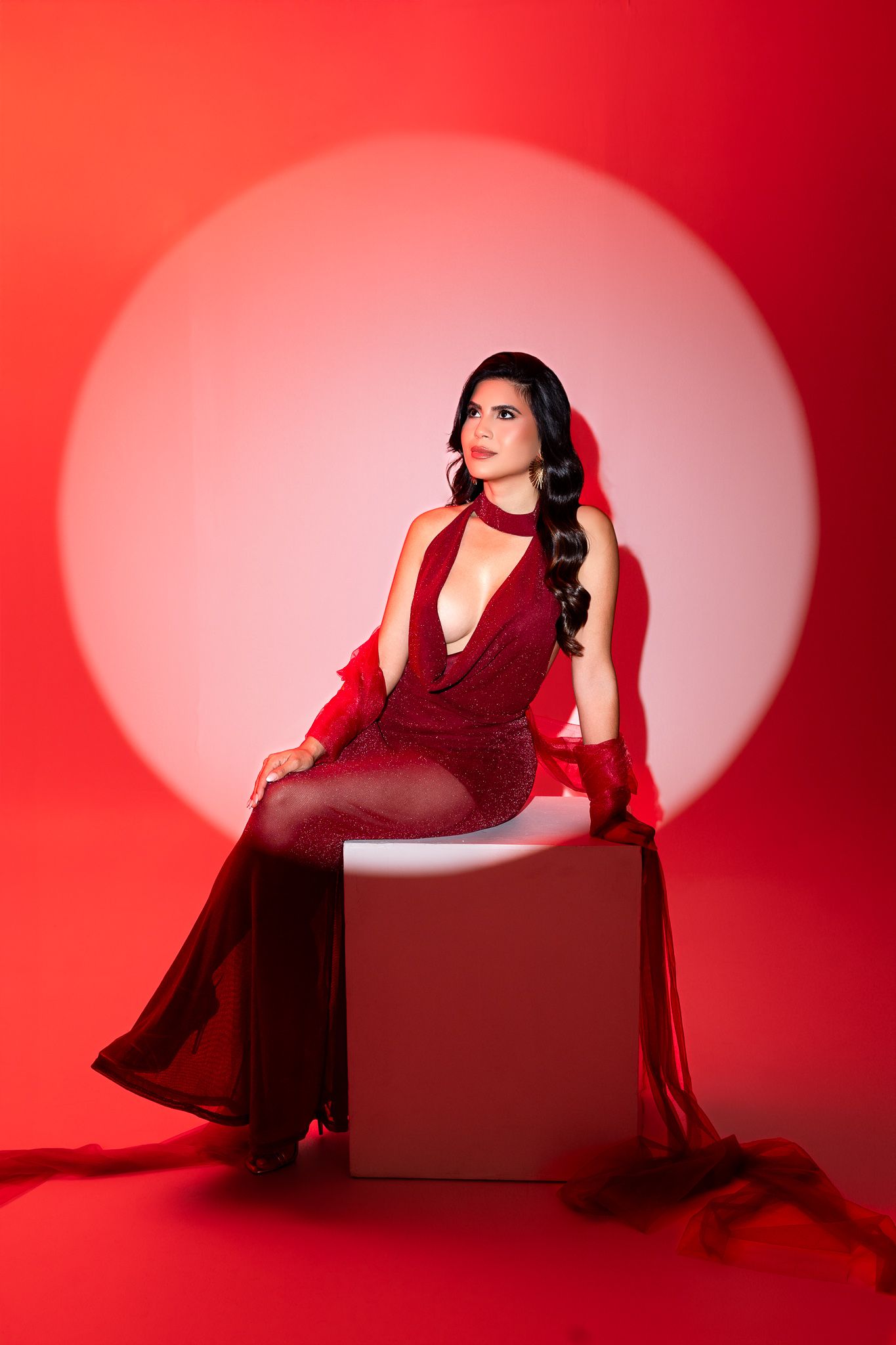 Figure in a glamorous red gown poses on a cube against a dramatic red backdrop with circular lighting effect.