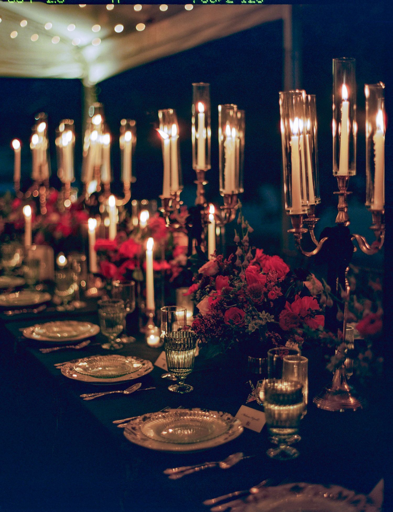Elegant table setting with tall candelabras, red floral arrangements, and gold-rimmed place settings for a formal dinner.