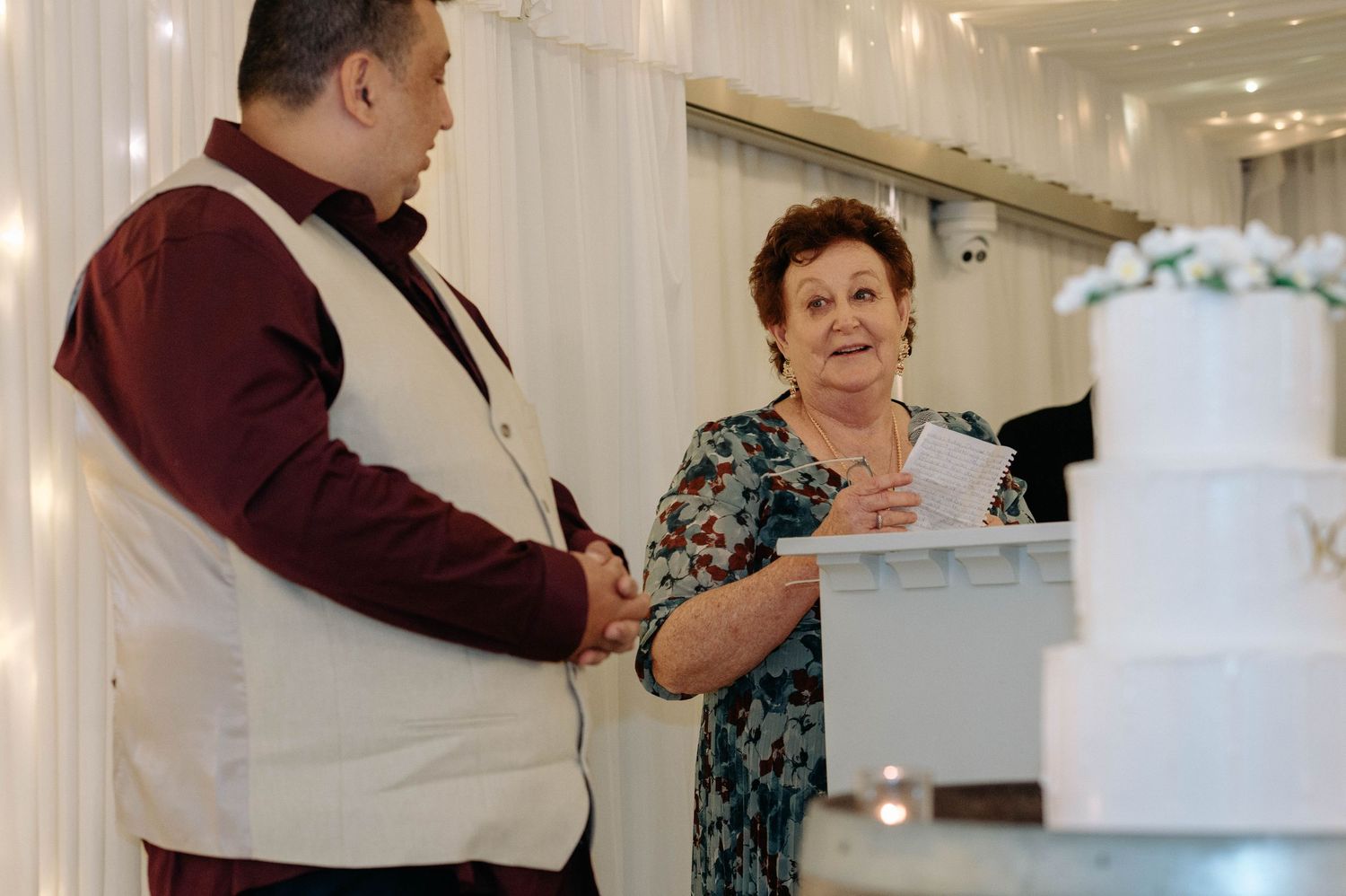 Two people stand at a counter discussing details while looking at an elegant white tiered cake on display.