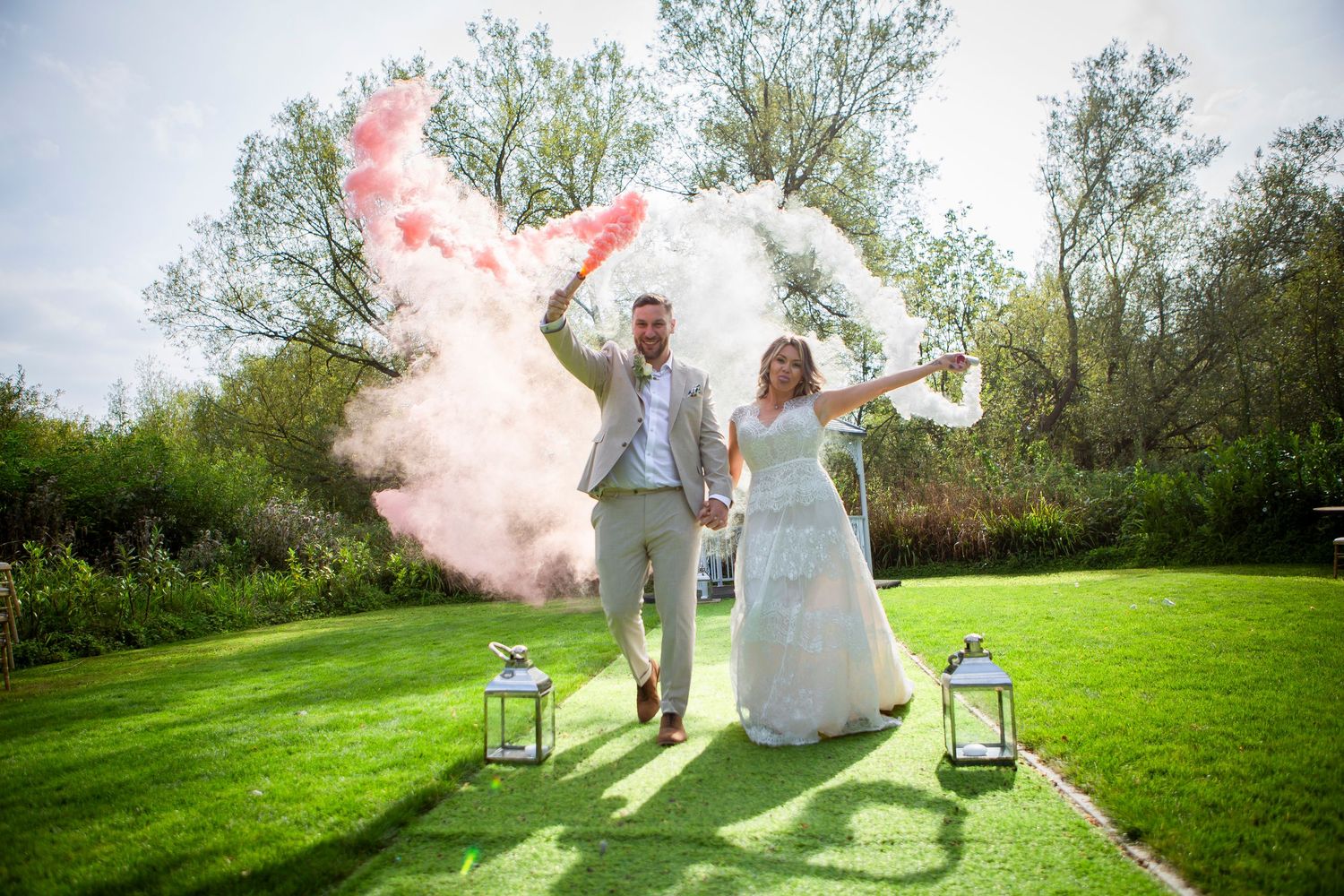 Bride and groom celebrate outdoors holding pink and white smoke bombs with lanterns on grass beside them.