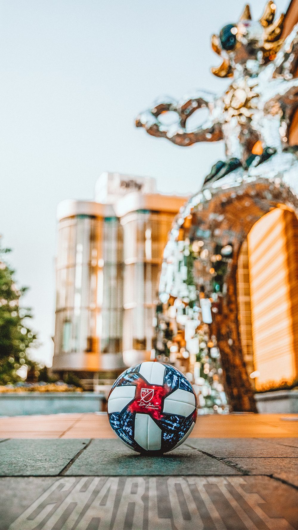 MLS soccer ball on a Charlotte city plaza with a decorative metallic sculpture and buildings in background.