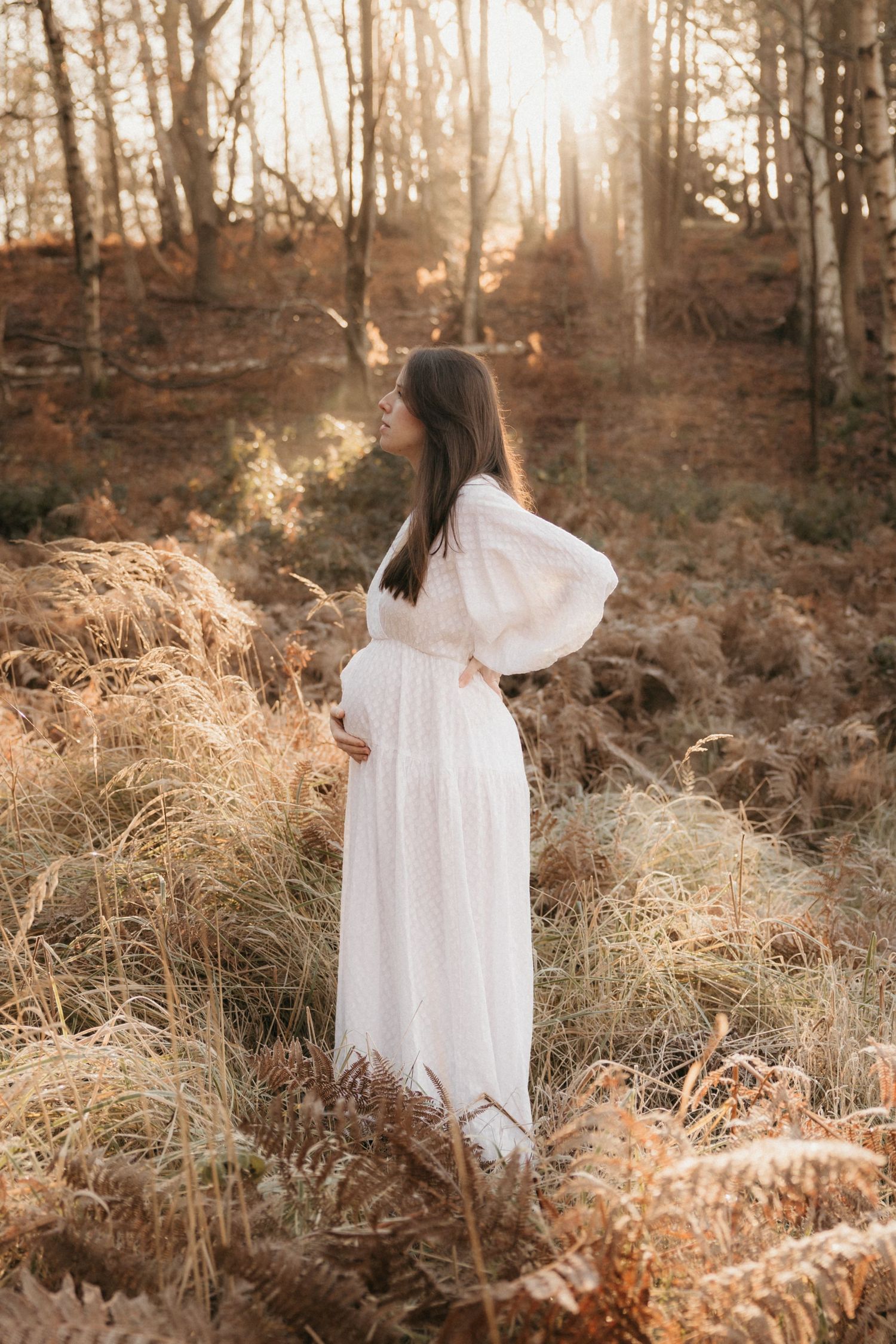 A couple sits together in a sunlit field, with gentle hands resting on a pregnant belly during maternity photoshoot.