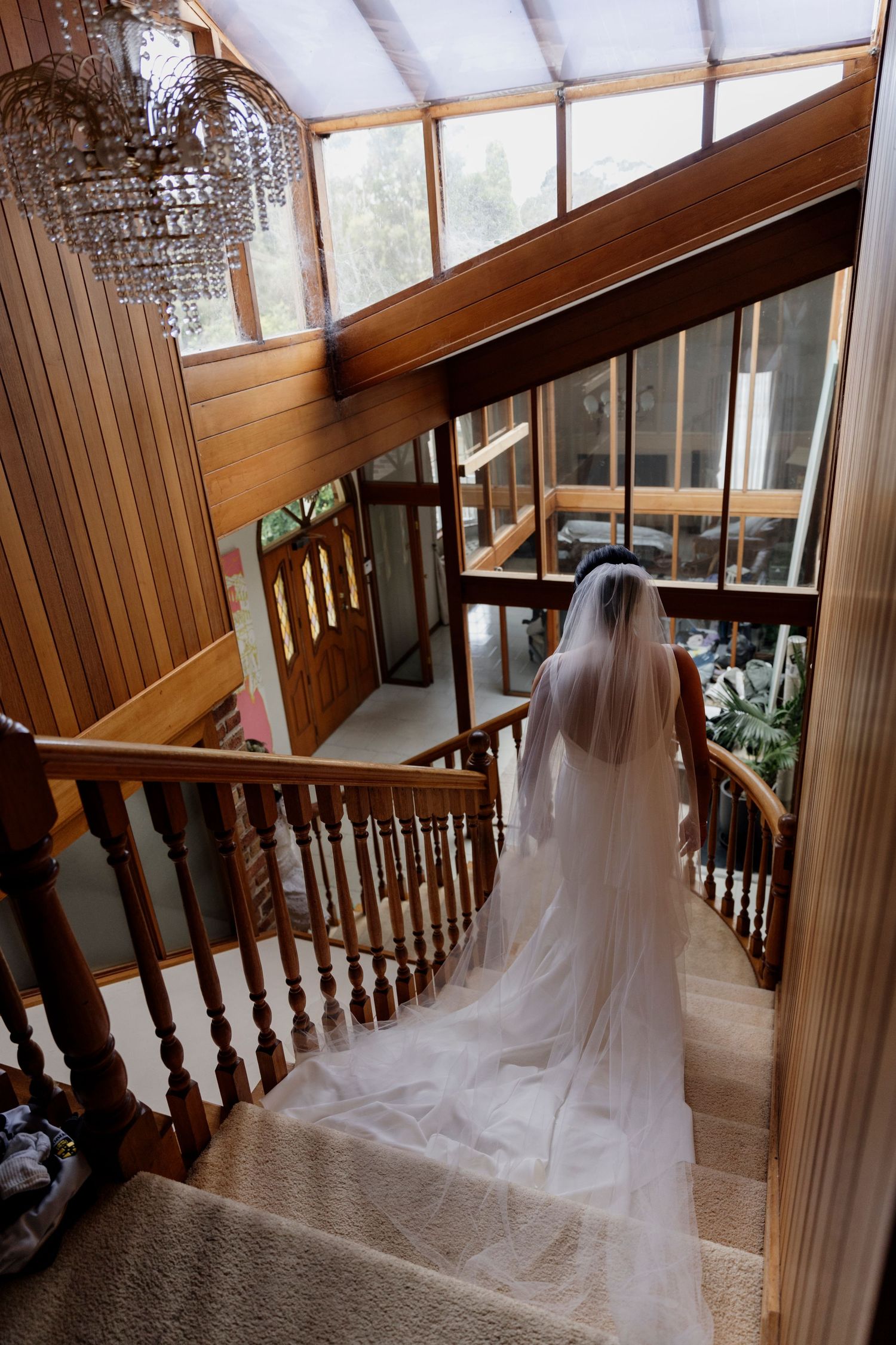 A flowing white wedding gown and long veil cascading down a wooden staircase with crystal chandelier and natural lighting.