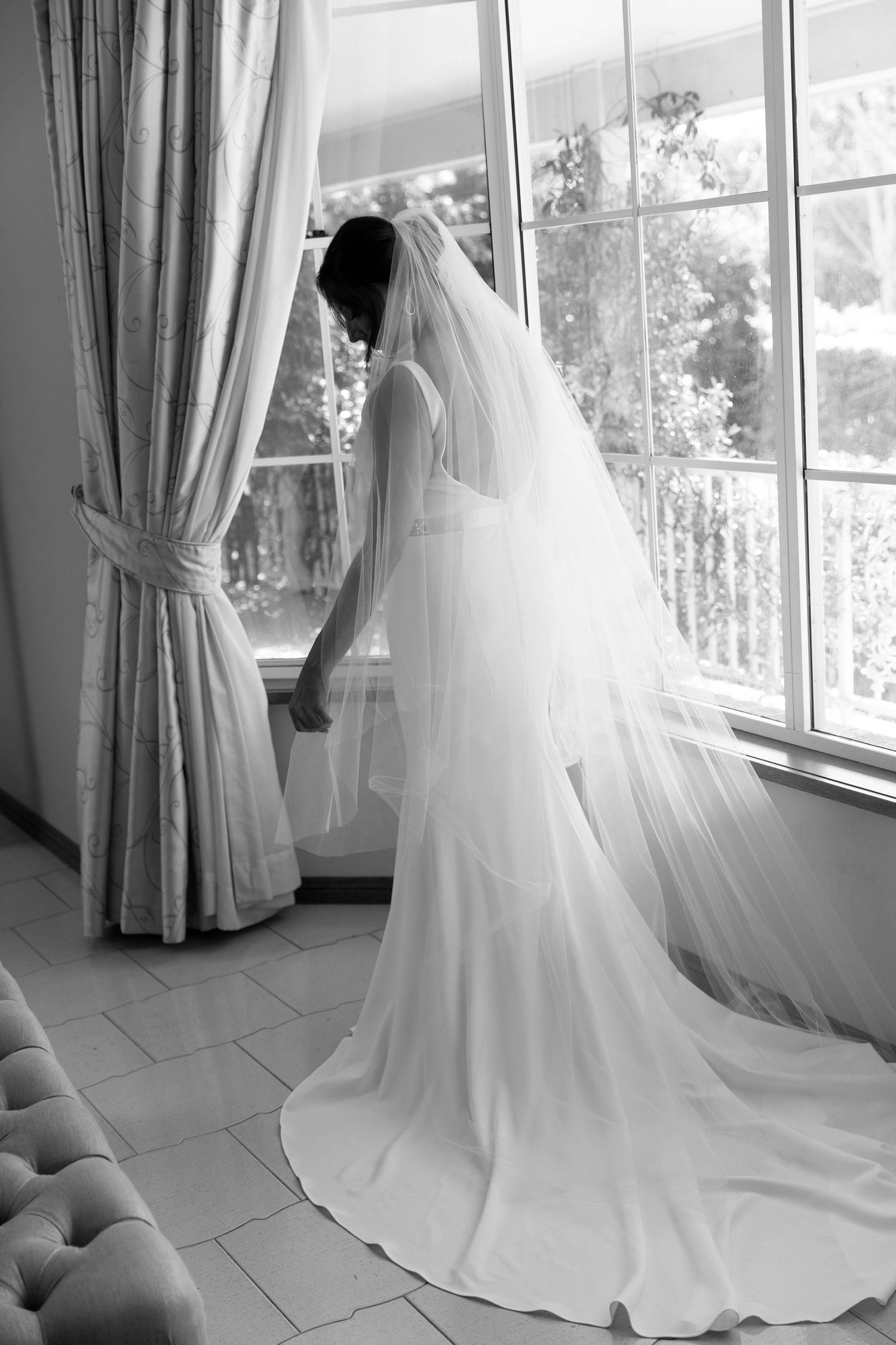 A bride in an elegant white gown and long flowing veil stands by a sunlit window with draped curtains.