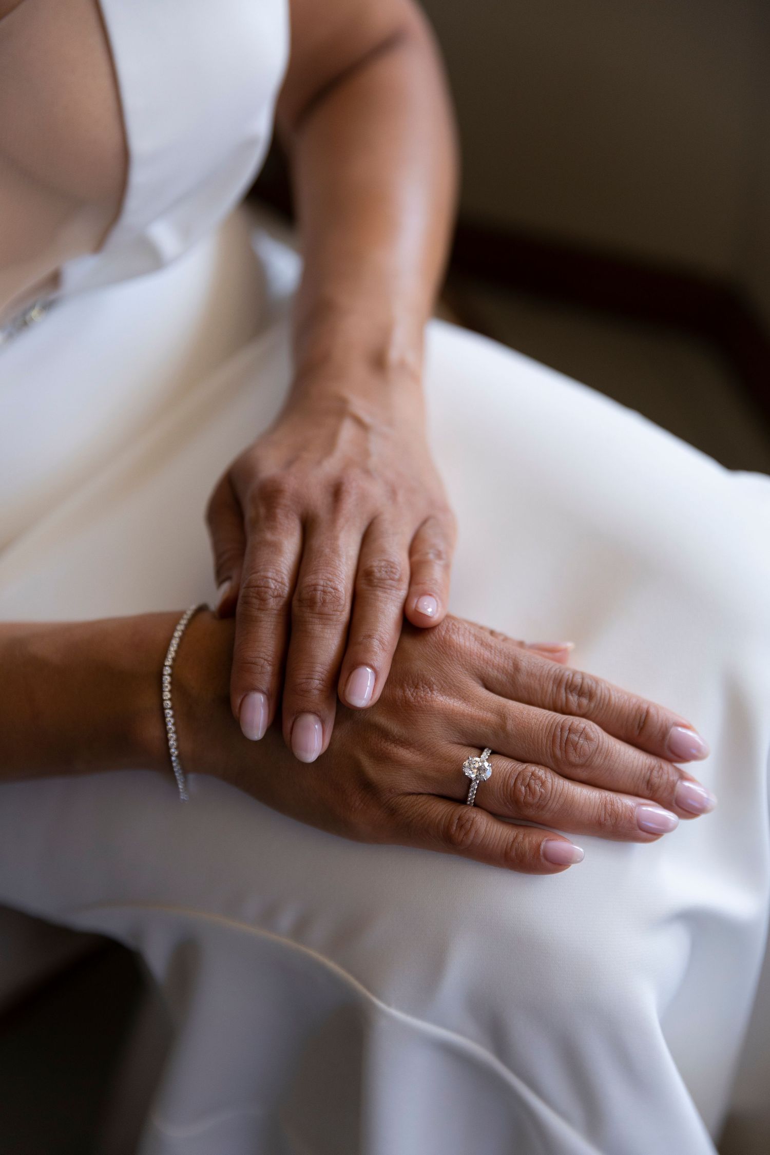 A close-up view of hands wearing an engagement ring resting on white fabric.