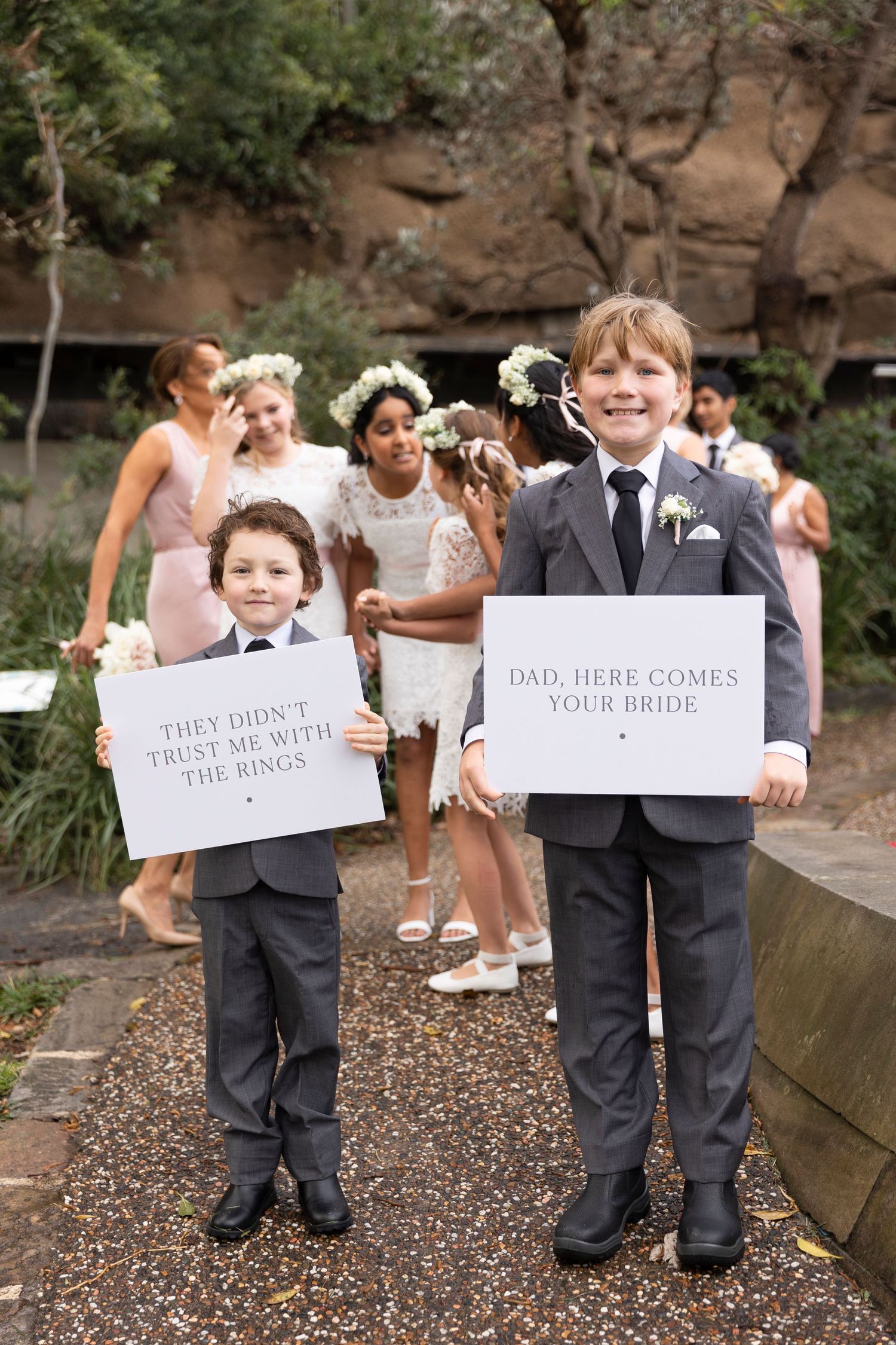 Two young ring bearers in suits hold wedding signs reading 'they didn't trust me with the rings' and 'here comes your bride' on a path.