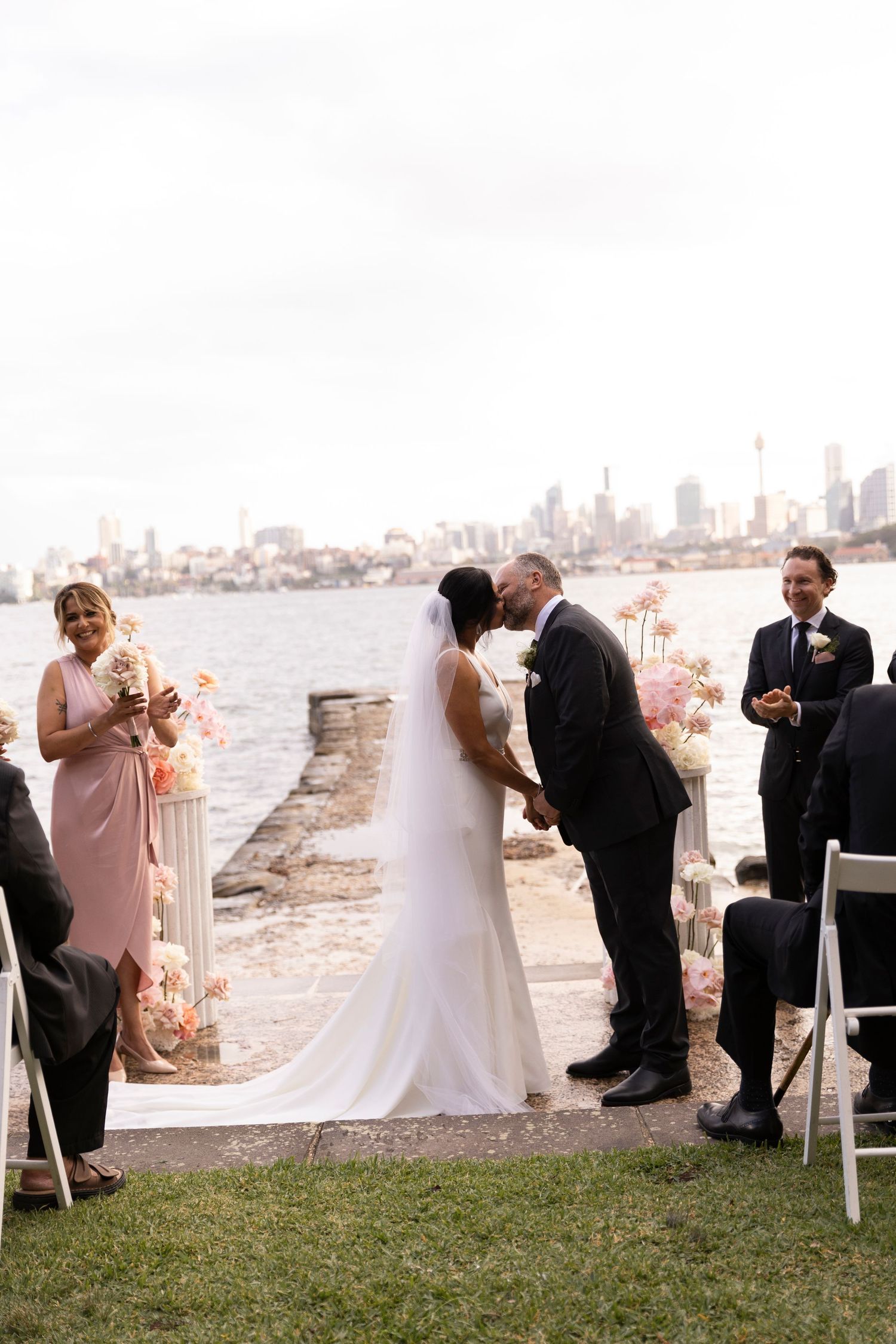 A waterfront wedding ceremony at sunset with the city skyline visible in the distance.