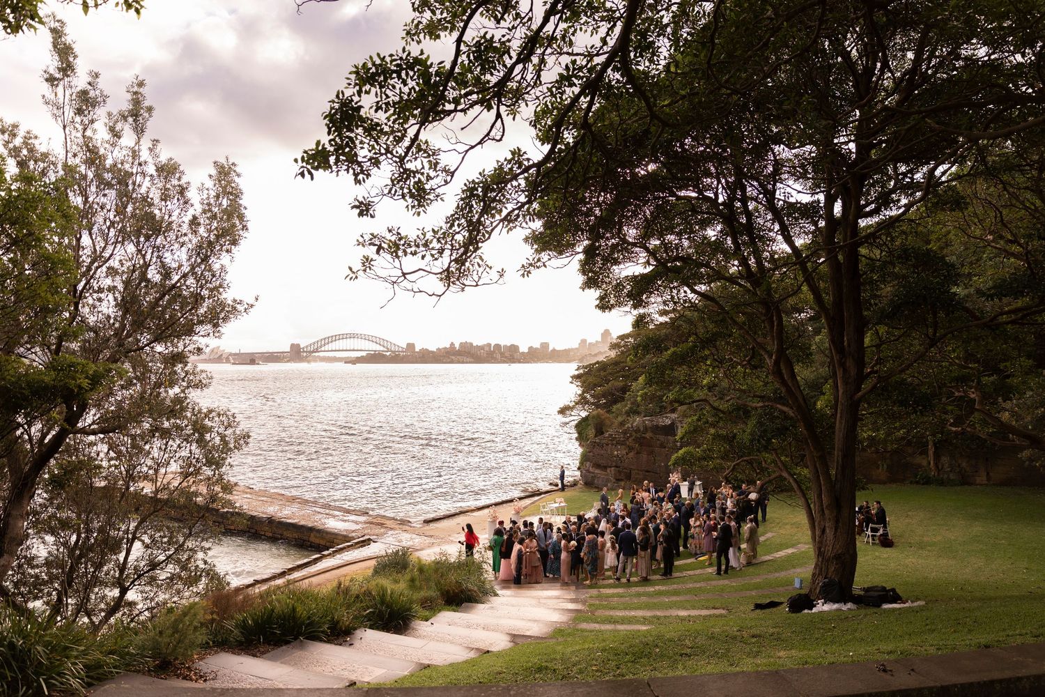 Wedding guests gathered on scenic coastal path with water and trees in background during outdoor ceremony.