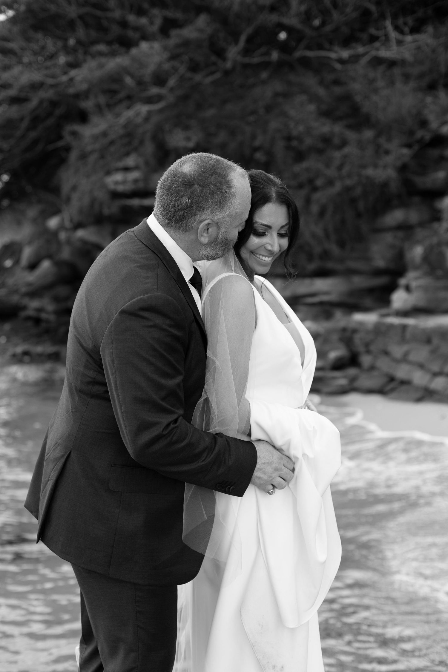 A happy couple in wedding attire shares a sweet embrace at the beach with ocean waves in the background.