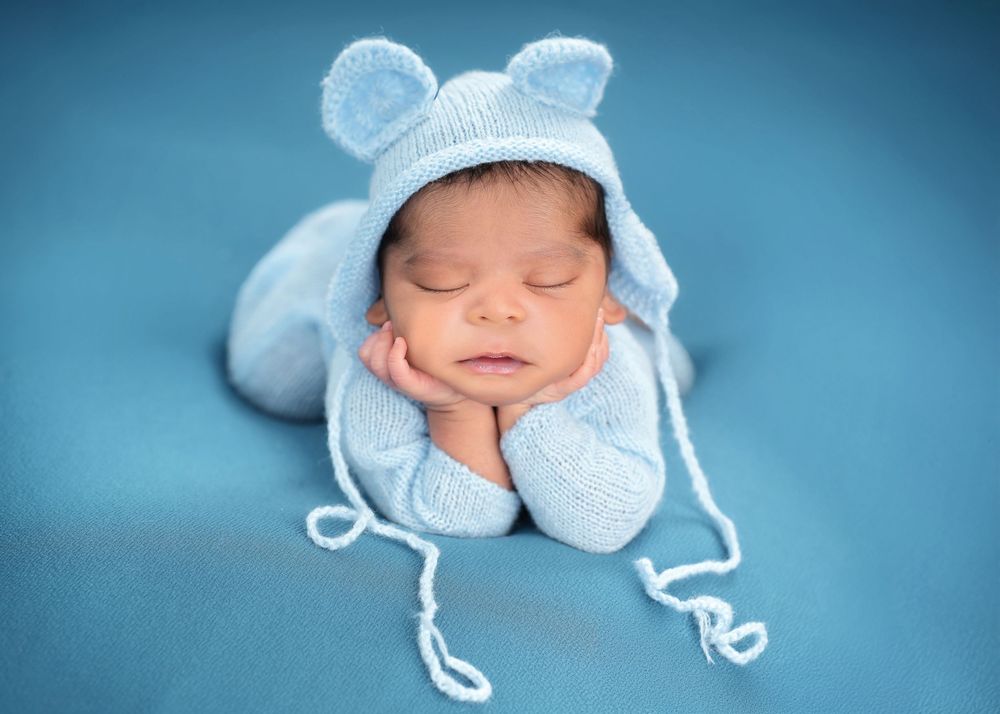 Sleeping newborn in light blue knitted bear hat laying on turquoise backdrop.
