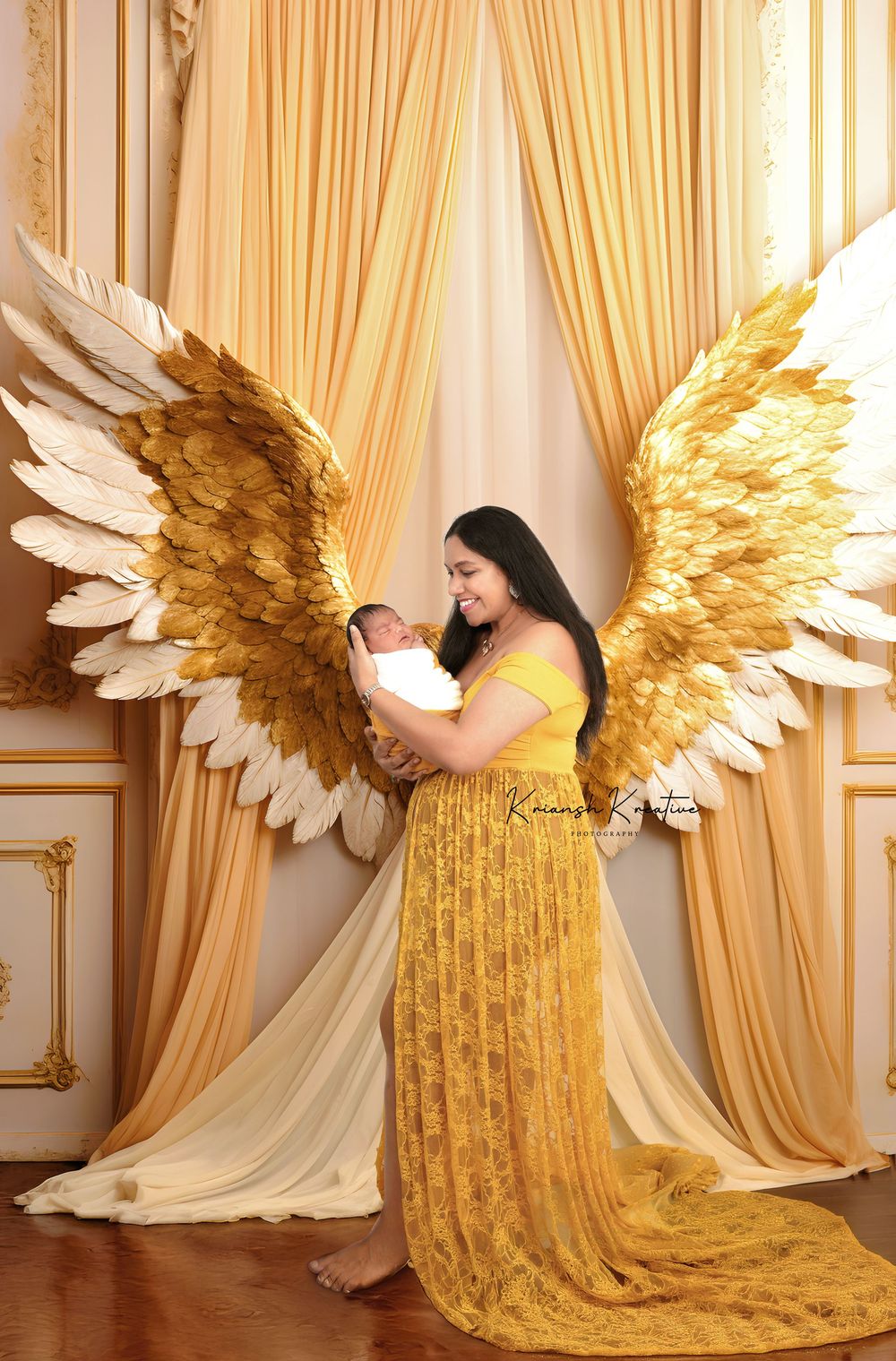 A woman in a flowing yellow lace gown poses with wings against gold curtains in an elegant studio setting.