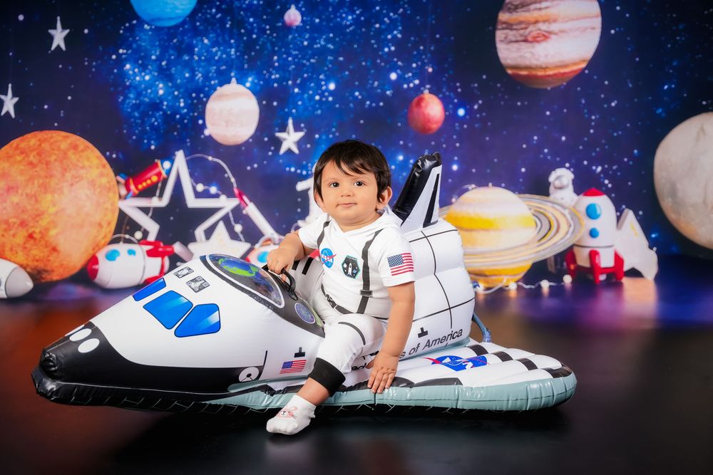 A space-themed photo with child in astronaut costume sitting on toy space shuttle with planets in background.