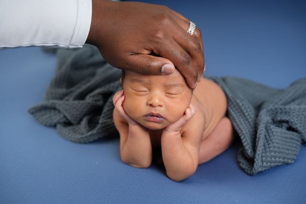 A gentle hand rests on a sleeping newborn baby positioned on a blue backdrop with a gray blanket.