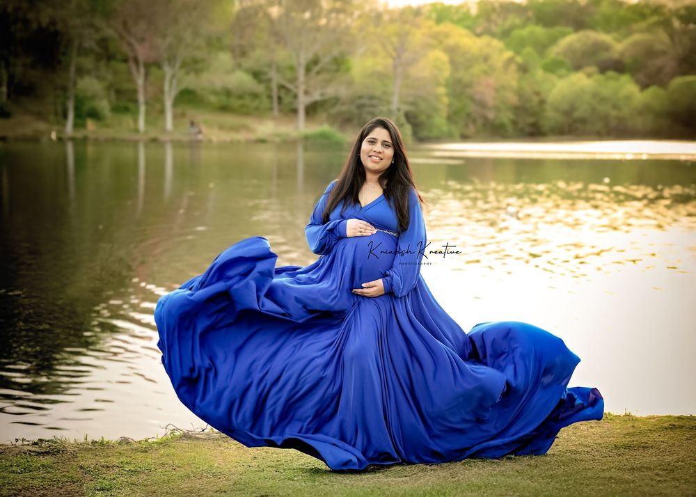 A flowing royal blue gown billows dramatically by a serene lake during golden hour at a tranquil park setting.