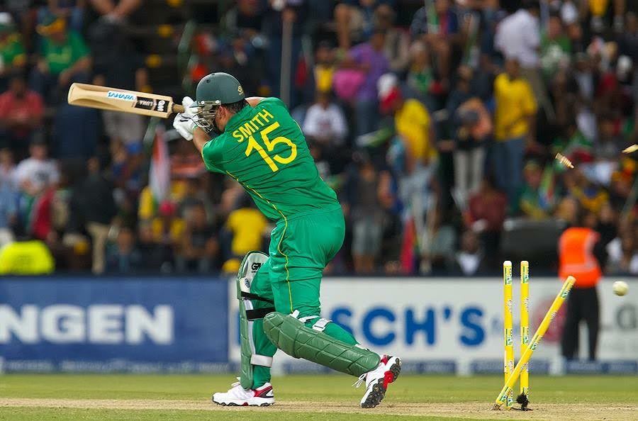 Cricket player in green uniform falls over during a match while attempting to play a shot as stumps get knocked over.