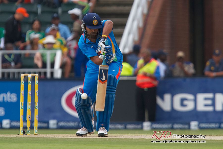 A cricket batter in blue uniform takes stance at the crease during a match on a professional field.