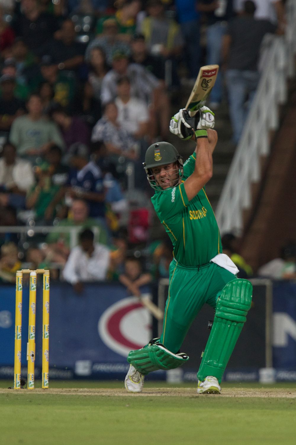 South African cricket batsman in green uniform plays an aggressive batting shot during a match at a stadium.
