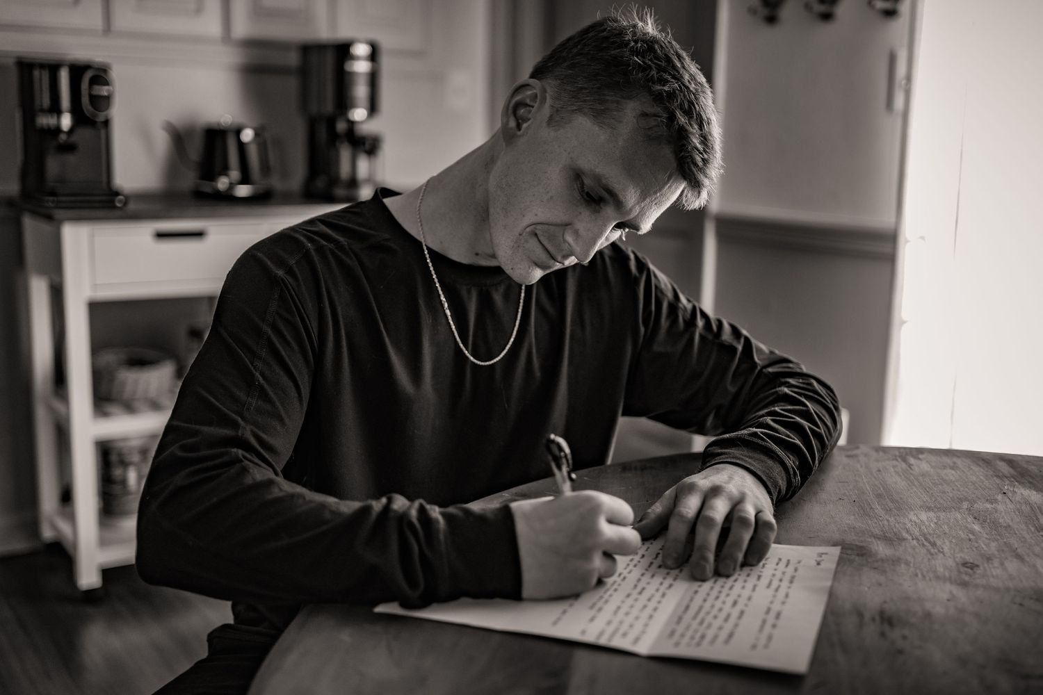 A person in a black sweater sits at a wooden table writing on paper in a dimly lit room.
