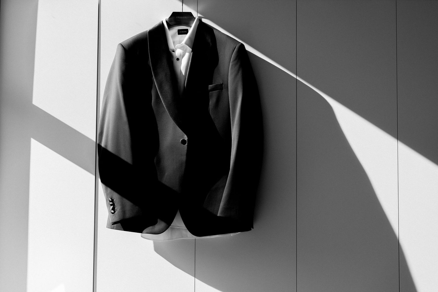 A black blazer suit jacket hanging on a white wall with dramatic diagonal shadows cast across it.